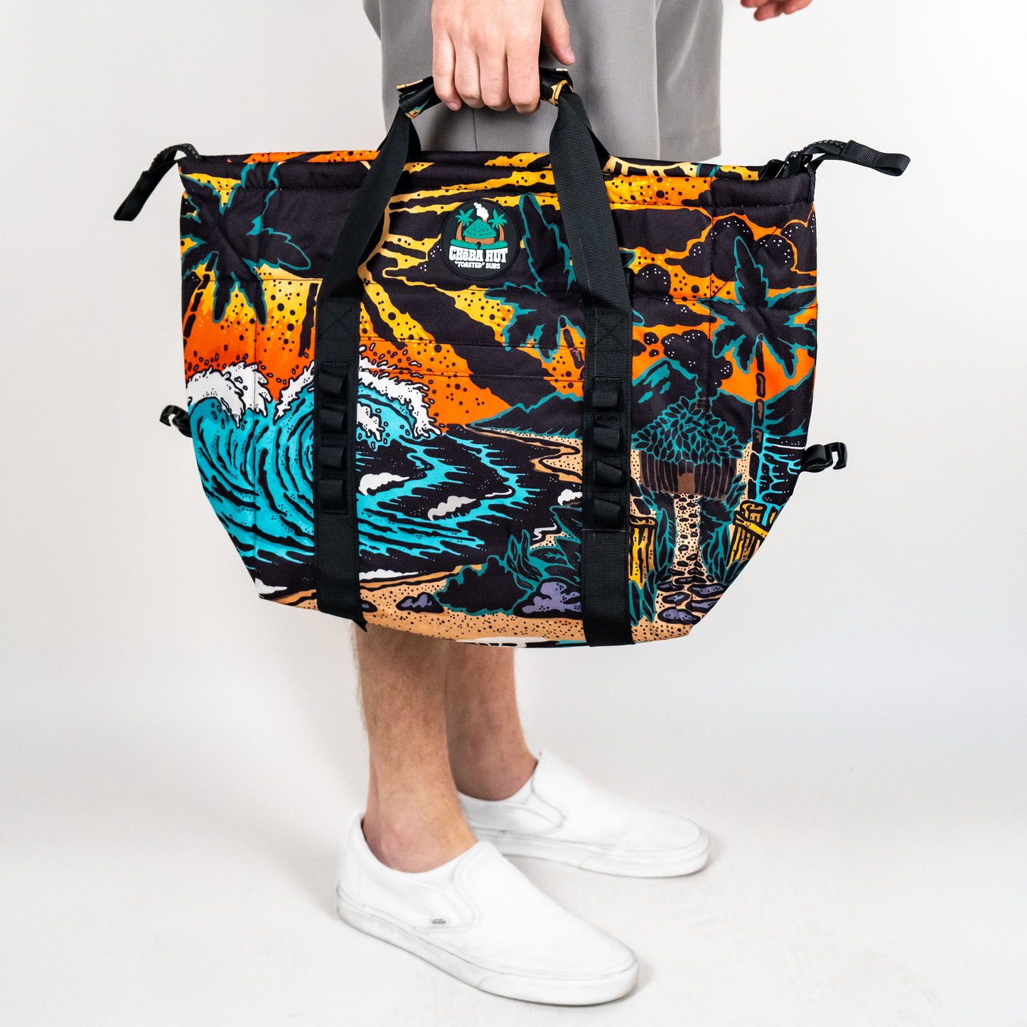 A person is shown from the knees down, holding a square-shaped cooler bag with both hands. The bag has a black fabric body with a colorful, detailed beach scene pattern featuring a large blue and white wave, a sandy beach, a dark blue ocean, an orange and yellow sunset sky, silhouetted palm trees, and a small hut. The person is wearing light gray pants and white slip-on shoes. The background is a plain light surface.