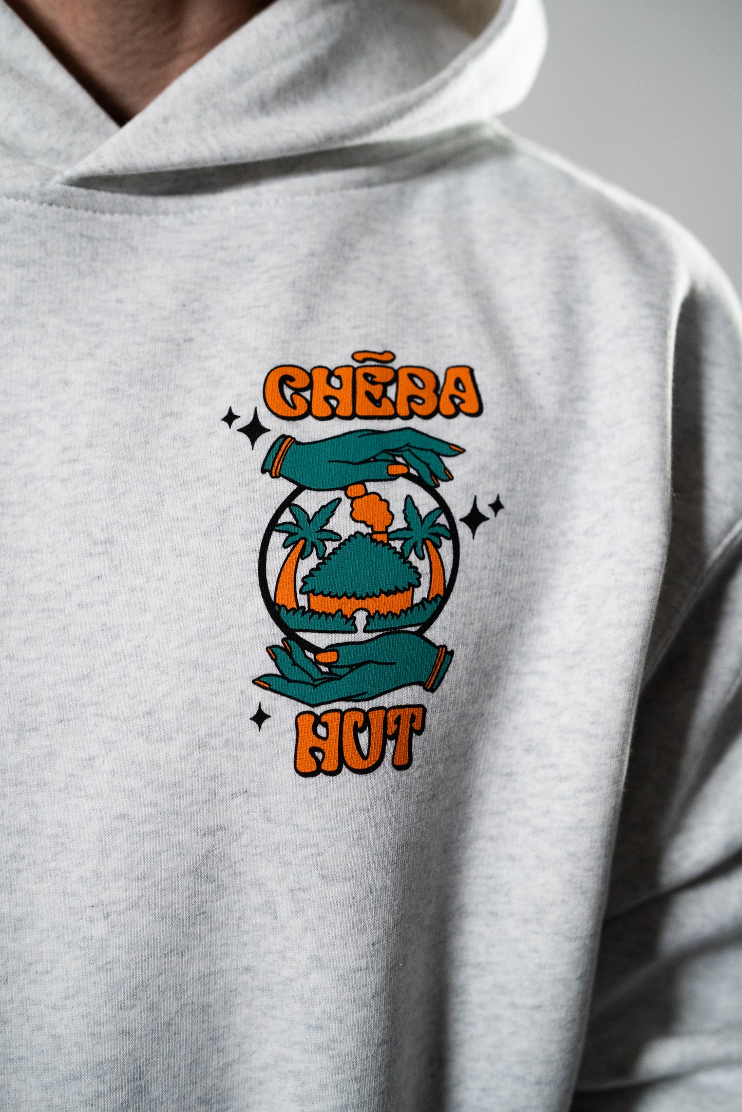 A person is wearing a light gray hooded sweatshirt with a graphic on the left chest featuring the 'Cheba Hut' logo with a globe and palm trees.