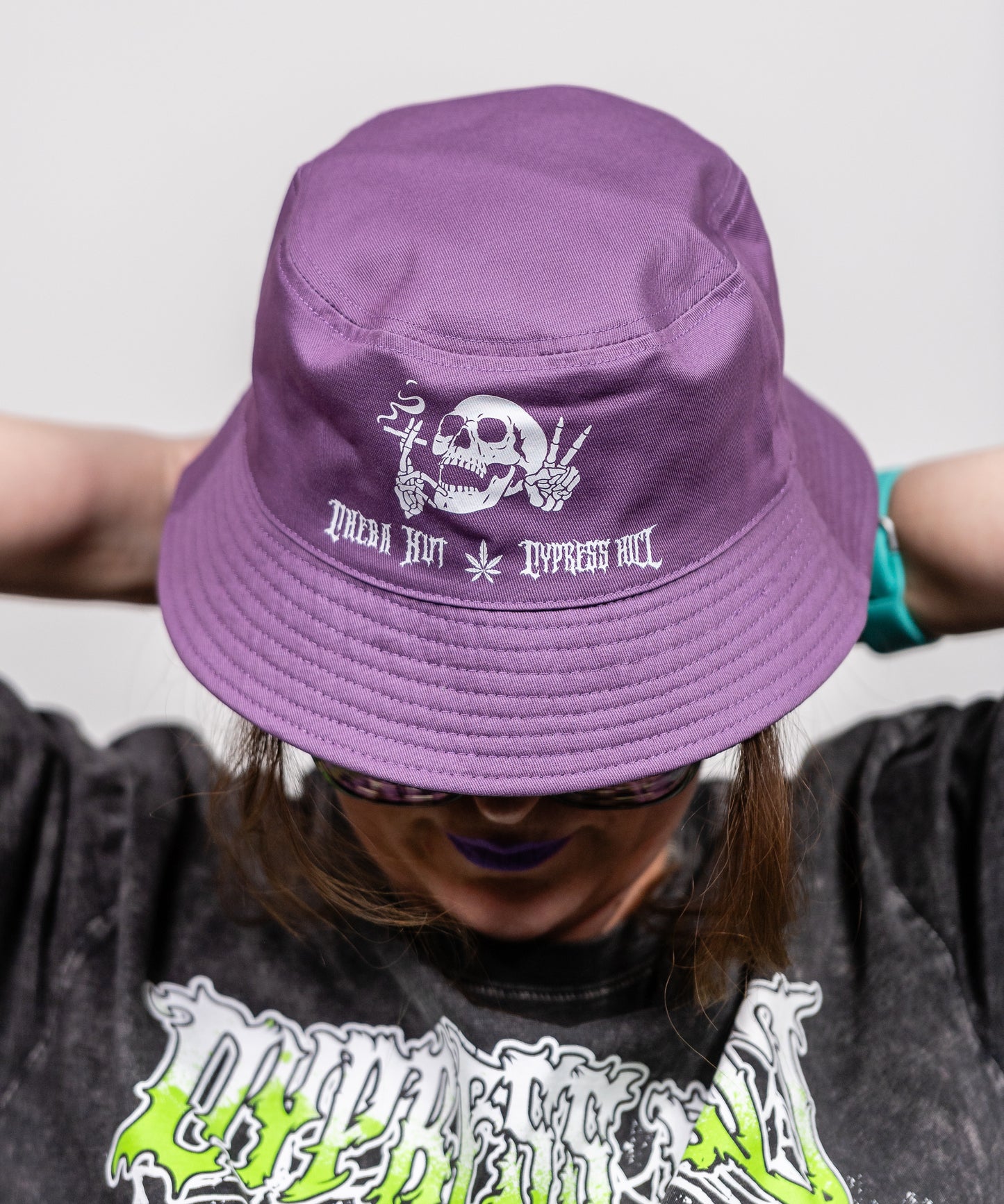 Image of a woman wearing a purple bucket hat. The front of the hat has a graphic of a skull smoking a joint and flashing a peace sign. Below the image are the words "Cheba Hut Cypress Hill".