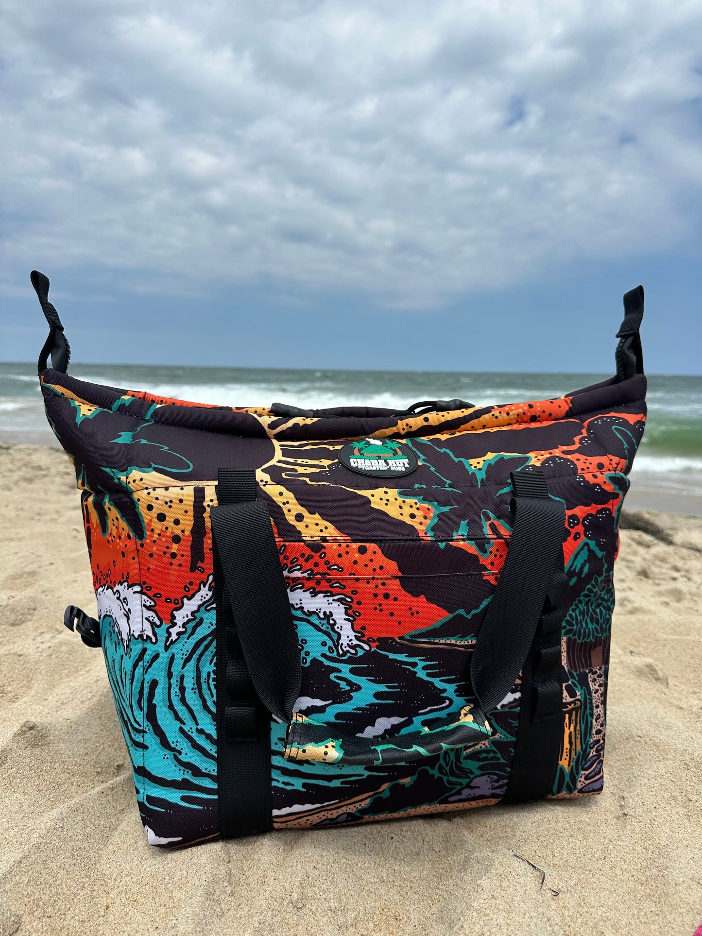 The bag has a black fabric body with a colorful, detailed beach scene pattern. The pattern includes a large blue and white wave, a sandy beach, a dark blue ocean, an orange and yellow sunset sky, silhouetted palm trees, and a small hut. The bag has black fabric handles, black zipper closures, and black fabric straps with buckles on the sides. The bag is placed on a sandy surface with a blue sky background and ocean waves.