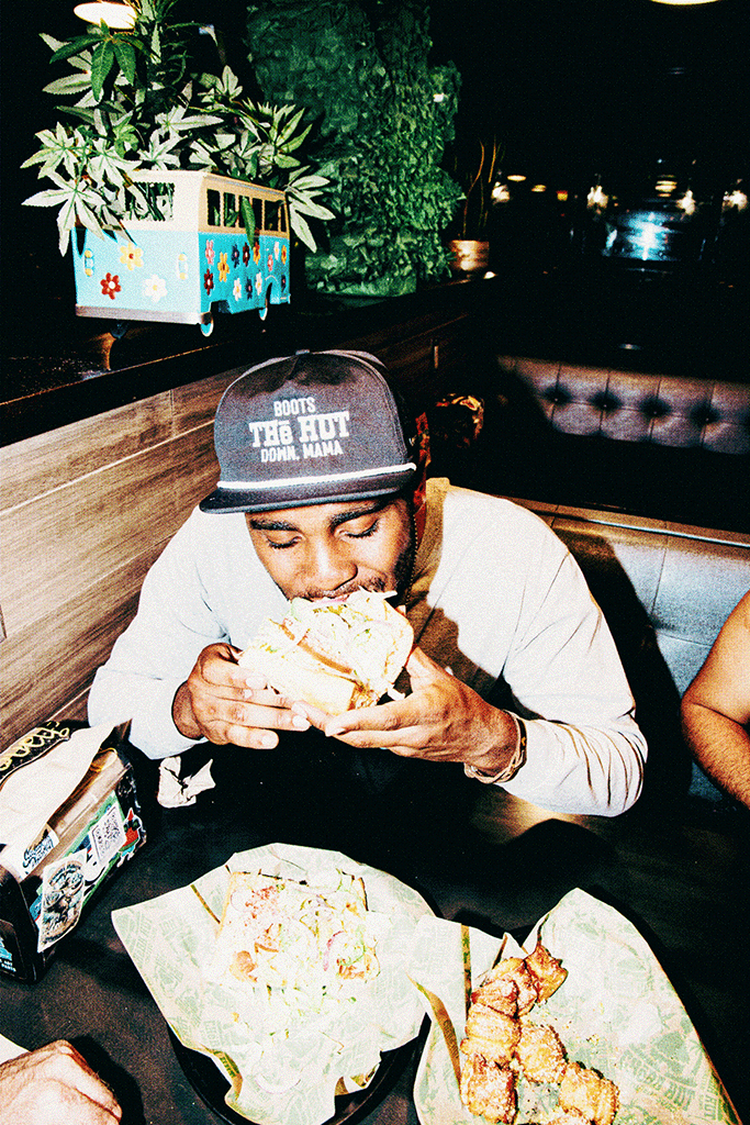 An image of a man with dark hair wearing a black flat billed hat. The hat has the words "Boots The Hut Down, Mama" embroidered in white thread on the front. The side of the hat also has the words "Cheba Hut Pride" embroidered in white thread.