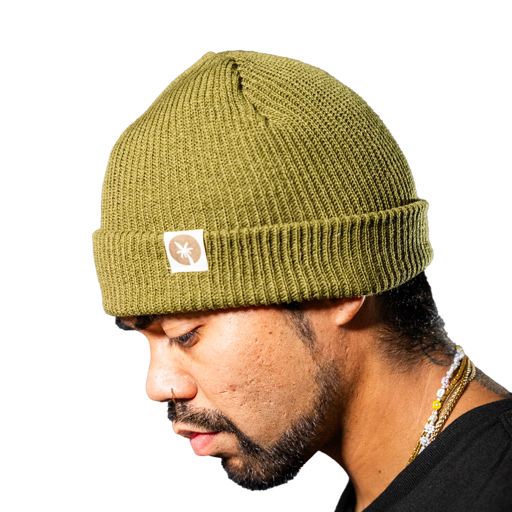 A close-up of a man wearing a green ribbed knit beanie with a small black and white palm tree logo patch on the side, set against a plain white background.