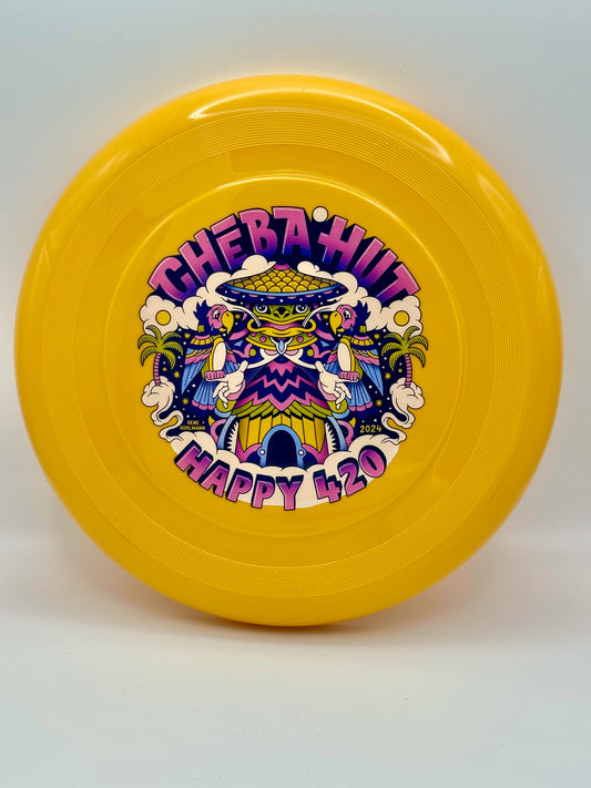 A bright yellow, nine-inch frisbee features a colorful circular graphic in the center. The graphic displays the words "CHEBA HUT" in stylized purple and pink lettering at the top, arching over a central image. This central image is a vibrant, cartoonish depiction of a building with a teal and purple roof, flanked by palm trees, clouds, and what appear to be parrots in shades of pink, blue, and yellow. Below the building, the words "HAPPY 420" are written in large, playful purple letters.