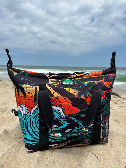 The bag has a black fabric body with a colorful, detailed beach scene pattern. The pattern includes a large blue and white wave, a sandy beach, a dark blue ocean, an orange and yellow sunset sky, silhouetted palm trees, and a small hut. The bag has black fabric handles, black zipper closures, and black fabric straps with buckles on the sides. The bag is placed on a sandy surface with a blue sky background and ocean waves.